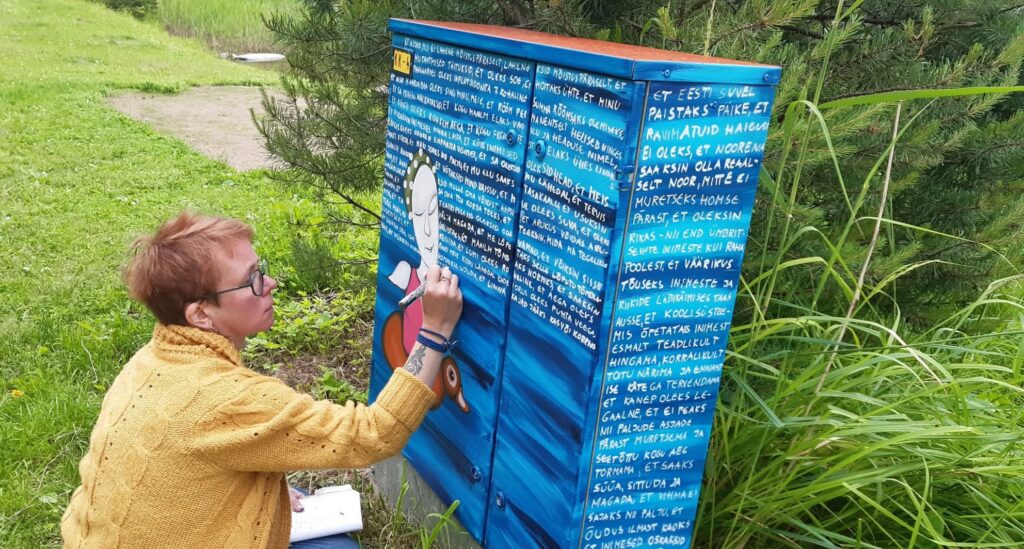 Naine kollases kampsunis maalib siniseks värvitud elektrikappi, kuhu on kirjutatud valge kirjaga eestikeelne tekst ja kujutatud stiliseeritud inimest. Taustal on rohelised puud ja muru.