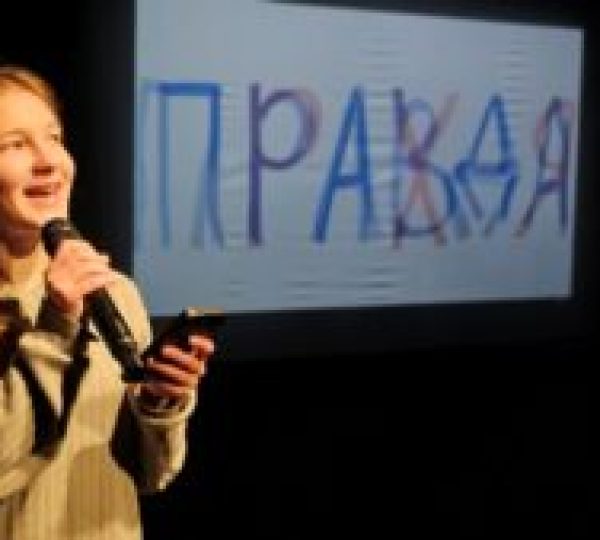 Tüdruk esineb mikrofoniga laval, taustal ekraanil venekeelne sõna „правда“ (tõde), mille peale on joonistatud mitmetähenduslikke ülekirjutusi.
